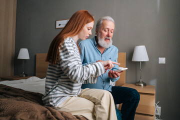 Handsome bearded senior grandfather learning to using mobile phone under guidance of pretty young granddaughter sitting on bed at home. Portrait of female teaching grandpa using smartphone.