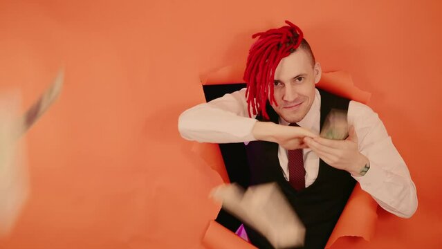 Close up of businessman hipster. Portrait man with red dreadlocks on his head throwing money banknotes, through a hole in the orange wall in the studio