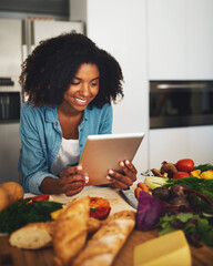 Looks like Im doing everything right. Shot of a cheerful young woman browsing on a digital tablet while being surrounded by vegetables in the kitchen at home during the day.