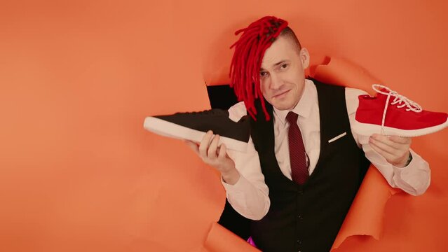 A smiling shoe salesman with red dreadlocks demonstrates trainers in a hole in the wall. Stylish man shows sneakers holding in his hands