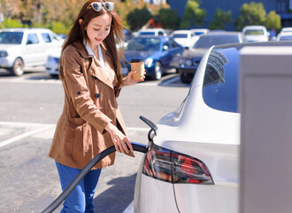 Smiling young woman standing on city parking near electric car, charging automobile battery from...