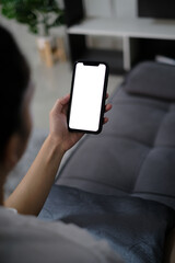 asia women holding smart phone with mockup white blank display, empty screen for app ads sitting on couch at home. Mobile applications technology concept, over shoulder close up view.