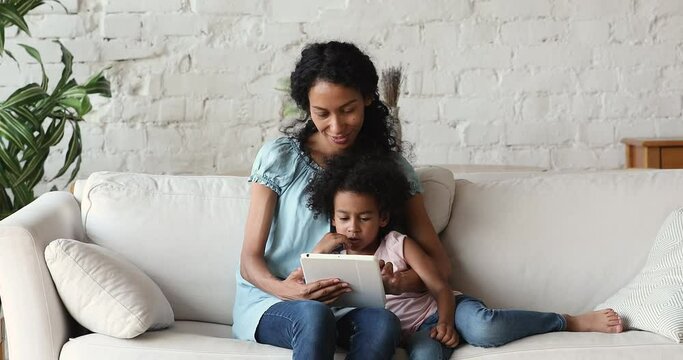 Millennial African American mother little child daughter hug on sofa at home enjoy playing videogame together using digital touchpad device. Happy mom with preschool age girl scroll pages on tablet pc