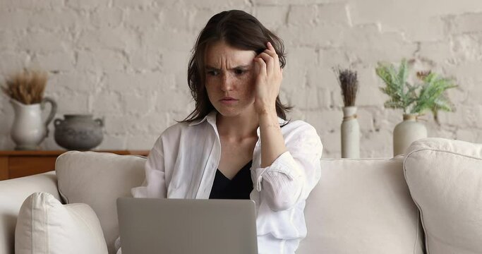 Angry teen female sit on couch look on laptop pc screen find problem in new app configuration irritated with low internet connection speed. Annoyed young woman get computer broken before saving data