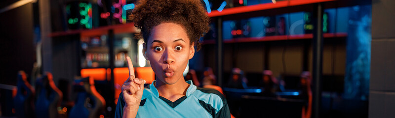 Cute African woman gamer in blue t-shirt pointing up by finger with emotional look in cyber club