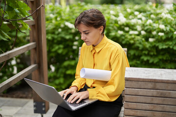 Fototapeta premium Business, finance industry or real estate concept. Successful smiling business lady in yellow blouse sitting on bench outdoor in park using laptop working or having online meeting. High quality image