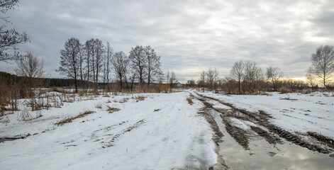 Broken dirt road with puddles in early spring. Dramatic winter landscape at sunset.