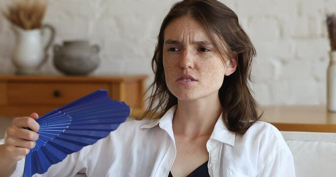 Sweaty exhausted teen female hold paper fan in hand wave herself suffer of too hot dry air uncomfortable climate indoor. Sad tired young woman struggle with heat at home feel bad without ac ventilator