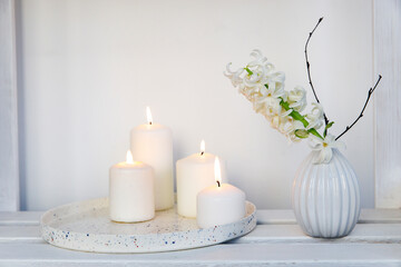 Fototapeta na wymiar A bouquet of white cut hyacinth a in a small white corrugated vase and three large burning candles on a round tray are on a beige table. Place for text