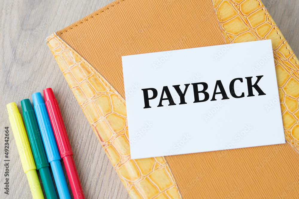 Poster payback text on a card on a notebook on a wooden table next to colored markers