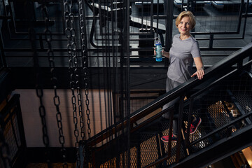 Obraz na płótnie Canvas Joyful woman with drink standing on stairs in gym