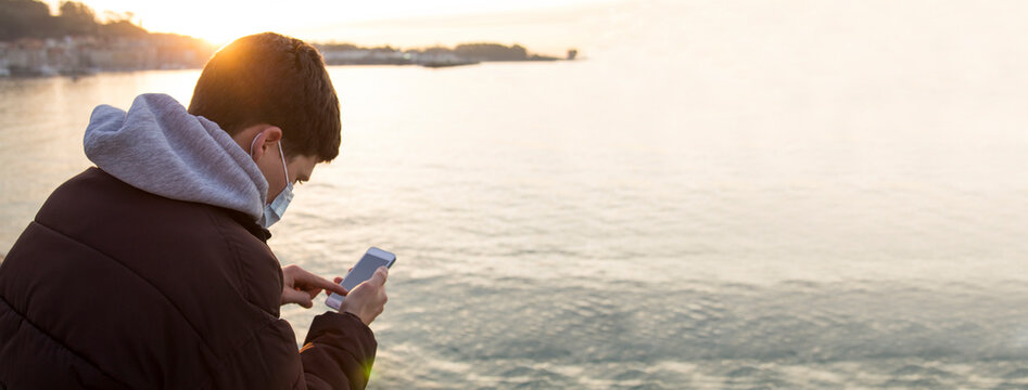 young man with medical mask on his face using mobile phone with the ocean in the background