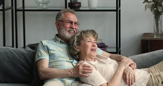 Attractive smiling carefree pensioner couple relax on sofa in living room, talking enjoy rest looking into distance. Real estate, comfort retired life, medical insurance for older gen citizen concept