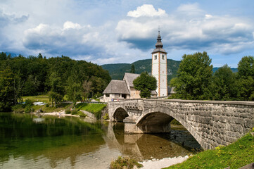 Church with a tall tower and an old stone bridge over the water at Lake Bohinj in Slovenia.
