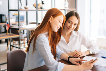 Side view of two creative young business women working using digital tablet at meeting desk with job documents at office. Business female colleagues using touchscreen computer for project discussion.