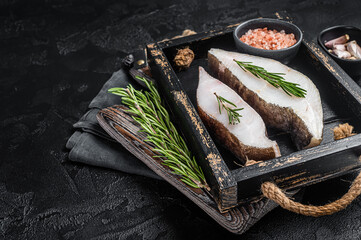 Raw halibut fish steak in wooden tray with herbs. Black background. Top view. Copy space