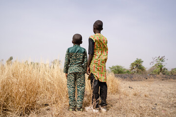 Rear view of two African boys standing in front of a barren field with tall dry grass staring at the horizon, questioning their fate; global warming, desertification and water scarcity concepts - obrazy, fototapety, plakaty