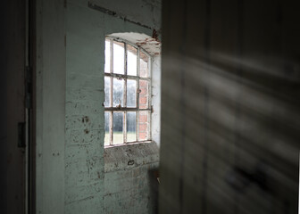 Dark and creepy wooden cellar window in abandoned workhouse viewed through open door bright sun light rays shining through on wall in scary sinister abandoned basement room. Atmospheric moody room.