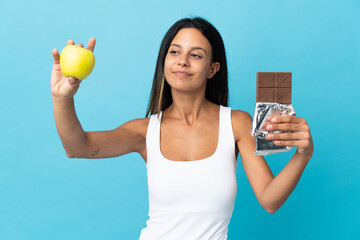 Caucasian girl isolated on blue background taking a chocolate tablet in one hand and an apple in...