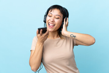 Young Uruguayan girl over isolated blue background listening music with a mobile and singing