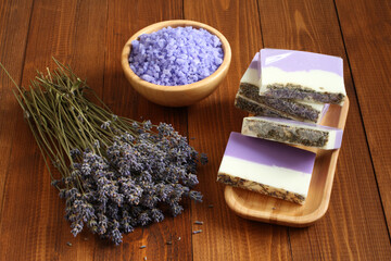 Sea salt with lavender in a bamboo bowl, soap made from lavender, olive oil and cocoa butter on a bamboo plate and dried lavender flowers are located on a brown wooden table. Closeup.