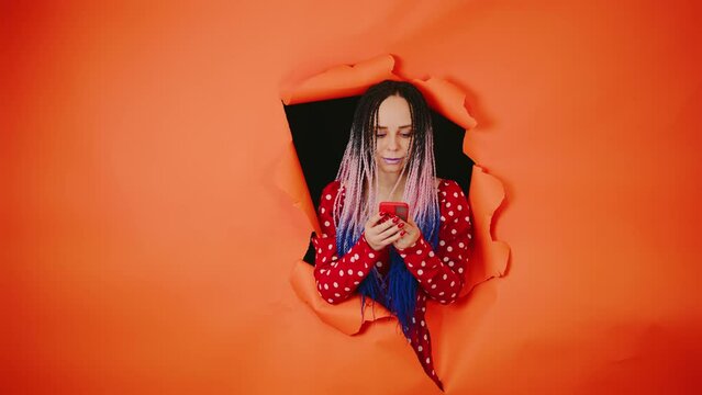 Bright woman browsing mobile phone and sticking out of hole of orange background. Impressive female with multicolored hairstyle using smartphone.