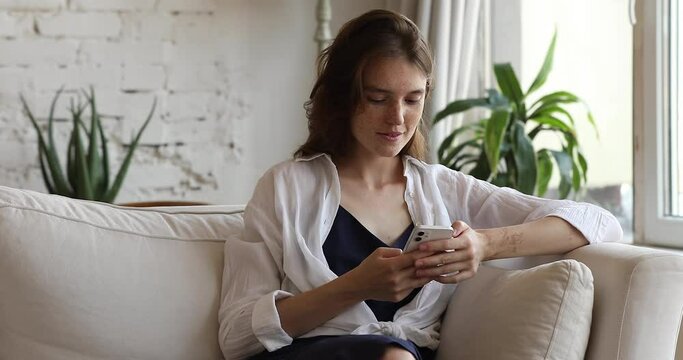 Pretty teen female sit on sofa at living room messaging with boyfriend in chat networking at social media using phone. Smiling young lady text answer on email play web game using wifi on smartphone
