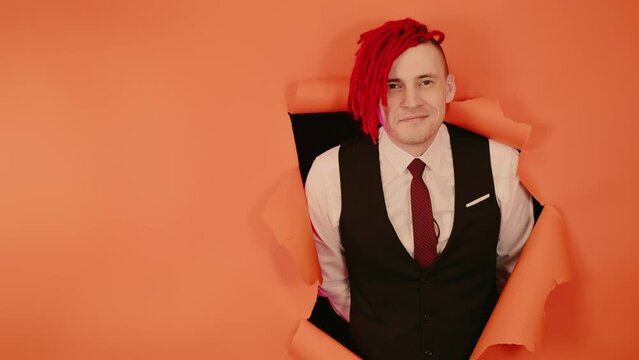 Smiling man with red dreadlocks in hole in wall. Stylish male with bright dreadlocks looking at camera through hole in orange wall in studio