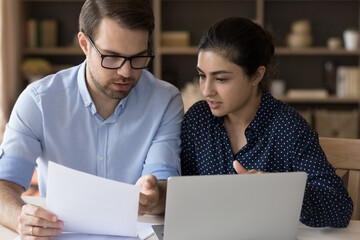 Serious multiethnic business partners coworkers students Indian woman Caucasian man sit at desk discuss document read sales report. Two young diverse teammates engaged in paperwork solve problem talk - Powered by Adobe