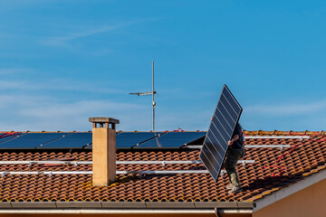 A technician hovering on a red tiled roof lifts a solar panel that he is about to add to a...