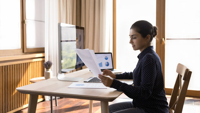 Focused Indian woman manager engaged in paperwork at home office do research using statistic information. Young lady marketing analyst study data in paper report on laptop and desktop computer screens