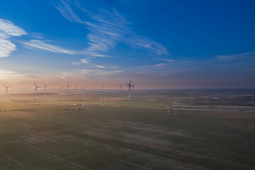 Rozległa równina pokryta polami uprawnymi i łąkami oświetlona światłem zachodzącego słońca. Na linii widnokręgu widać turbiny wiatrowe.