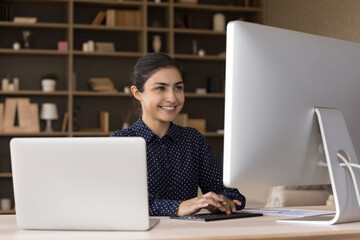 Successful young Indian female office employee executive work at desk using two computers print...