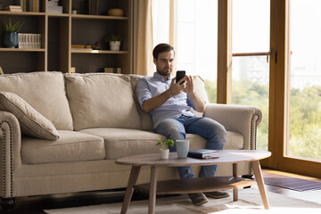Obraz premium Focused millennial man relax on sofa at living room listen to music in phone audio player app play online game wearing wireless headphones. Young guy in earphones watch video on cell screen on weekend
