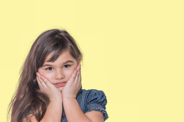 Tanned long haired little girl wears  denim costume with both hands on her face is posing on yellow background 