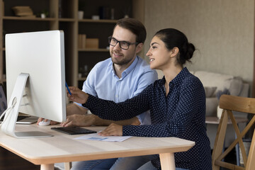 Young male in glasses trainee apprentice take professional internship work by desktop computer in team with experienced Indian female mentor. Two colleagues of diverse gender race interacting using pc