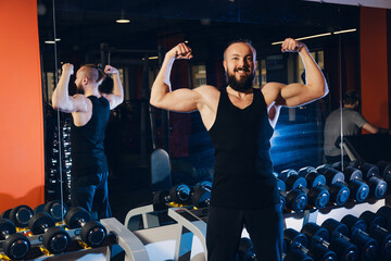 A man of athletic build poses against the background of a rack with dumbbells in the gym