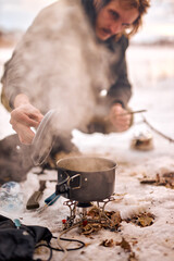 Fototapeta na wymiar young caucasian guy preparing food in winter forest. Bushcraft, hike, camping, travel, adventure and people concept. smoke from prepared cooked meal on small saucepan. in frozen
