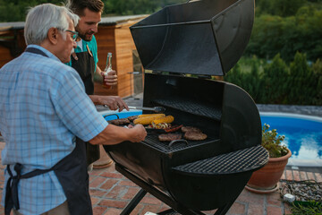 Senior man and son drinking beer and roasting meat and corn on barbecue in the backyard near the...