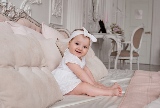 a smiling baby girl in a white headband and bodysuit is sitting on a bed in a room with a beautiful interior