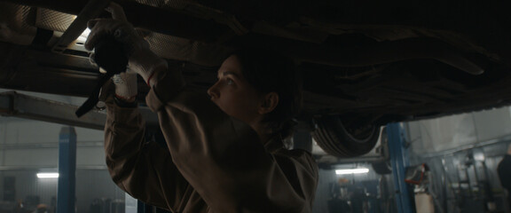 CU portrait of Caucasian female mechanic repairing a car in a workshop, working under car bottom. Shot with 2x anamorphic lens