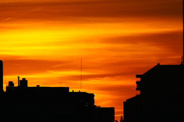 Fototapeta premium Dramatic cloudy sky at sunset in urban area. Silhouette buildings with the sun on yellow and orange background