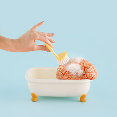 Young female hand with brush washes model of human brain in tub with foam on isolated pastel blue background. Minimal abstract concept of mental health, brain fog healthcare treatment or brainwashing.