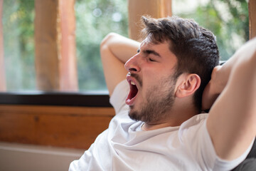 jeune homme assis dans un fauteuil qui s'étire en baillant. Il se repose car il est fatigué. On...