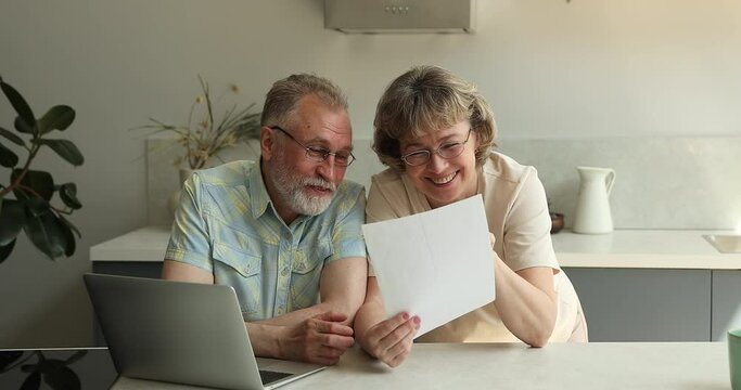 Older couple smile read good news in document, formal letter, talk sit at table indoors, discuss family budget, feel satisfied with money refund, plan investment, check bank savings statement concept