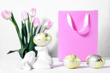 Easter is a bright holiday, the hare is holding an egg on his feet, there is a gift bag for sales and discounts nearby. Delicate pink spring flowers tulips. Yellow golden eggs in the marble technique