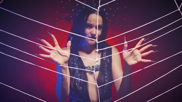 fantasy gothic vampire woman touches web with claws, looks in camera. Black sexy creative dress. Girl dark queen crown on head. halloween costume concept. Night cobweb scenery, smoke neon red light.