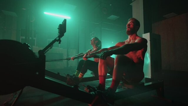 Muscular man and sporty woman workout on training simulator in crossfit gym. Muscular sporty man and women workout on training simulator in gym. Sportswomen exercising on rowing machines