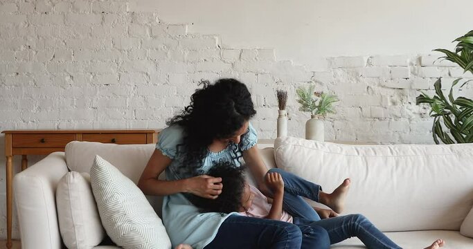 Happy laughing millennial African mother play with active energetic child tickle little girl on comfy sofa at living room. Overjoyed mama preschool daughter enjoy kidding messing at home giggle cuddle