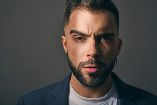 Male model shot in studio. Serious and confident expression. Angry or upset look and a casual style. Headshot of man with a beard, brown hair, brown eyes on a grey solid background. 